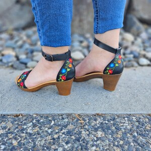 May include: Black leather heeled sandals with colorful floral embroidery. The sandals have a closed toe and an ankle strap.