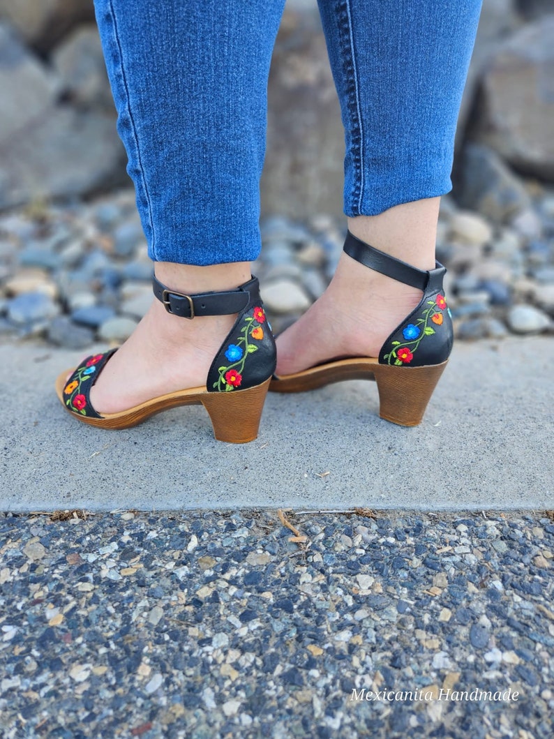 May include: Black leather heeled sandals with colorful floral embroidery. The sandals have a closed toe and an ankle strap.