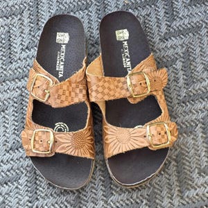 Op de afbeelding: Een paar beige leren sandalen met twee verstelbare bandjes met goudkleurige gespen. De bandjes zijn voorzien van een reli&euml;f met een zonnebloem- en ruitpatroon. Het donkerbruine voetbed bevat de witte tekst MEXICANITA HANDMADE.