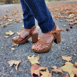 May include: Brown leather woven platform sandals with a chunky heel. The sandals are worn by a person wearing blue jeans and standing on a paved surface with fallen autumn leaves.