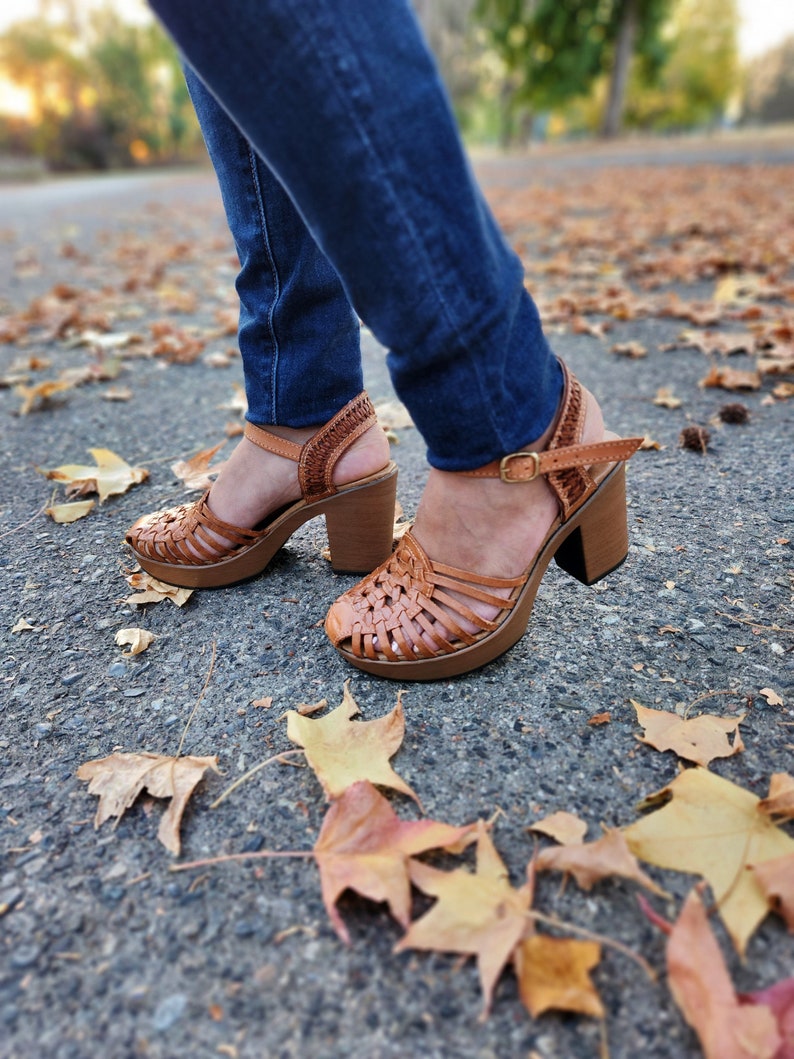 May include: Brown leather woven platform sandals with a chunky heel. The sandals are worn by a person wearing blue jeans and standing on a paved surface with fallen autumn leaves.