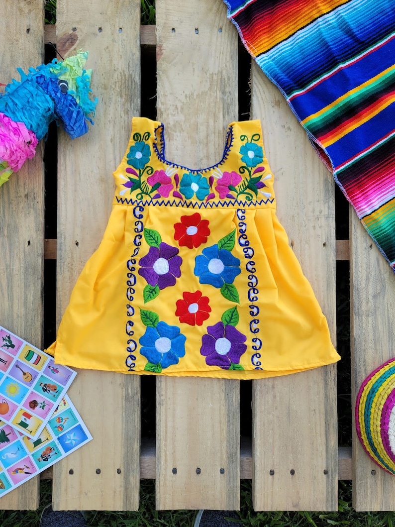 Pode incluir: Um vestido amarelo sem mangas com flores bordadas coloridas. O vestido tem uma bainha festoneada e uma borda azul ao redor do decote.