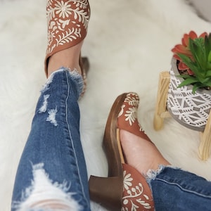 May include: Brown leather heeled shoes with white floral embroidery. The shoes have a wooden heel and are paired with distressed blue jeans. The shoes are a stylish footwear option.