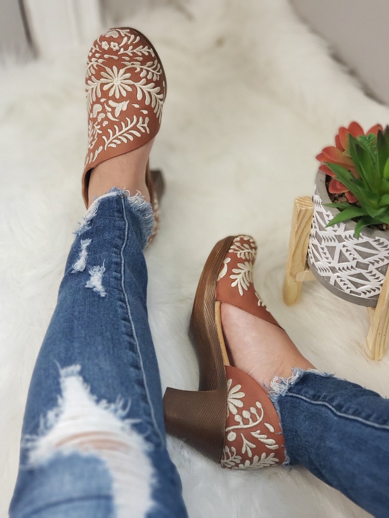 May include: Brown leather heeled shoes with white floral embroidery. The shoes have a wooden heel and are paired with distressed blue jeans. The shoes are a stylish footwear option.