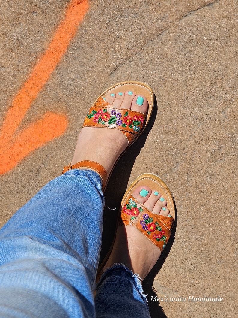 Pode incluir: Um par de sand&aacute;lias de couro marrom com bordados florais e esmalte de unha azul turquesa. As sand&aacute;lias s&atilde;o usadas por uma pessoa que est&aacute; vestindo jeans azul. O texto "Mexicanita Handmade" &eacute; vis&iacute;vel no canto inferior direito da imagem.