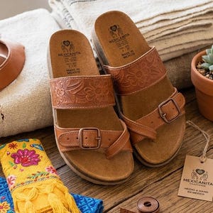 Pu&ograve; includere: Sandali in pelle marrone con cinturini goffrati a fiori e chiusure con fibbia. I sandali sono su una superficie di legno, con un'etichetta "Mexicanita". Sono presenti anche un metro a nastro arrotolato e un tessuto decorativo.