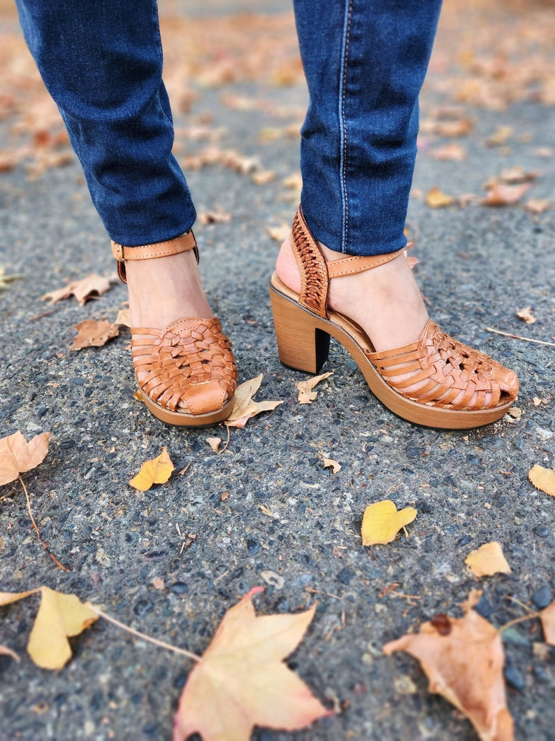 May include: Brown leather woven sandals with a chunky wooden heel. The sandals have an ankle strap and are worn with blue jeans.