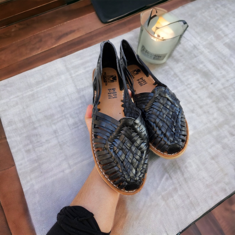 Puede incluir: Un par de sandalias de cuero negro con tiras tejidas. Las sandalias tienen una plantilla de cuero marr&oacute;n y un dedo cerrado.