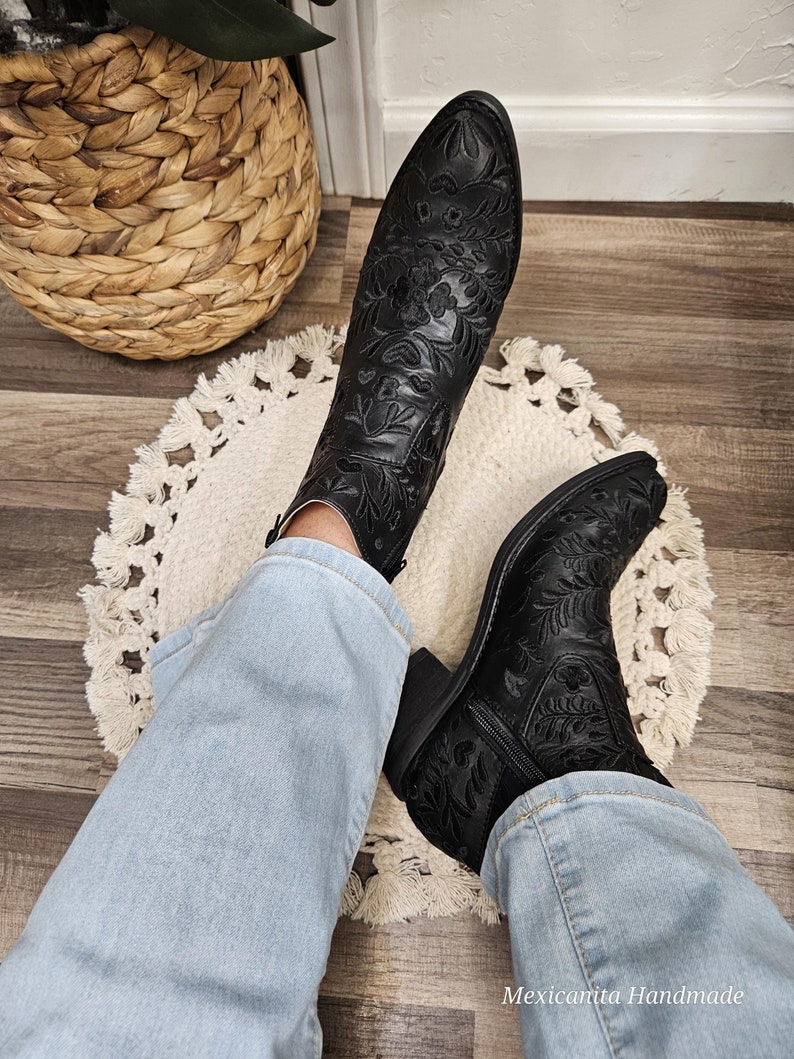 May include: Black ankle boots with floral embroidery. The boots have a pointed toe and a low heel. The boots are worn with blue jeans.