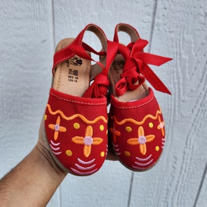 Puede incluir: Sandalias de lona rojas con flores bordadas y lazos de cinta roja. Las sandalias tienen una etiqueta de talla que dice "MEX 19 USA 12".