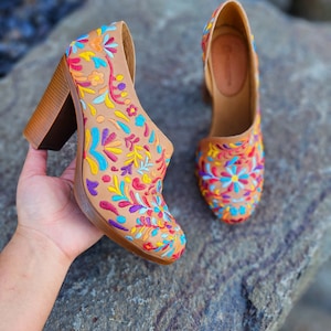 May include: A pair of brown leather heeled shoes with colorful floral embroidery. The shoes have a block heel and a pointed toe.