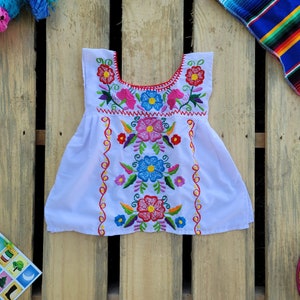 Pode incluir: Vestido branco bordado com desenhos florais coloridos. O vestido tem uma bainha vermelha ao redor do decote e uma borda festoneada.