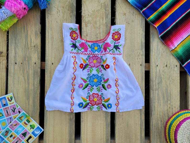Pode incluir: Vestido branco bordado com desenhos florais coloridos. O vestido tem uma bainha vermelha ao redor do decote e uma borda festoneada.