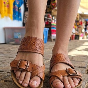 Pu&ograve; includere: Sandali in pelle marrone con dettagli floreali in rilievo sui cinturini. I sandali hanno un plantare in sughero e fibbie regolabili. L'immagine mostra i sandali ai piedi di una persona, con uno sfondo sfocato.