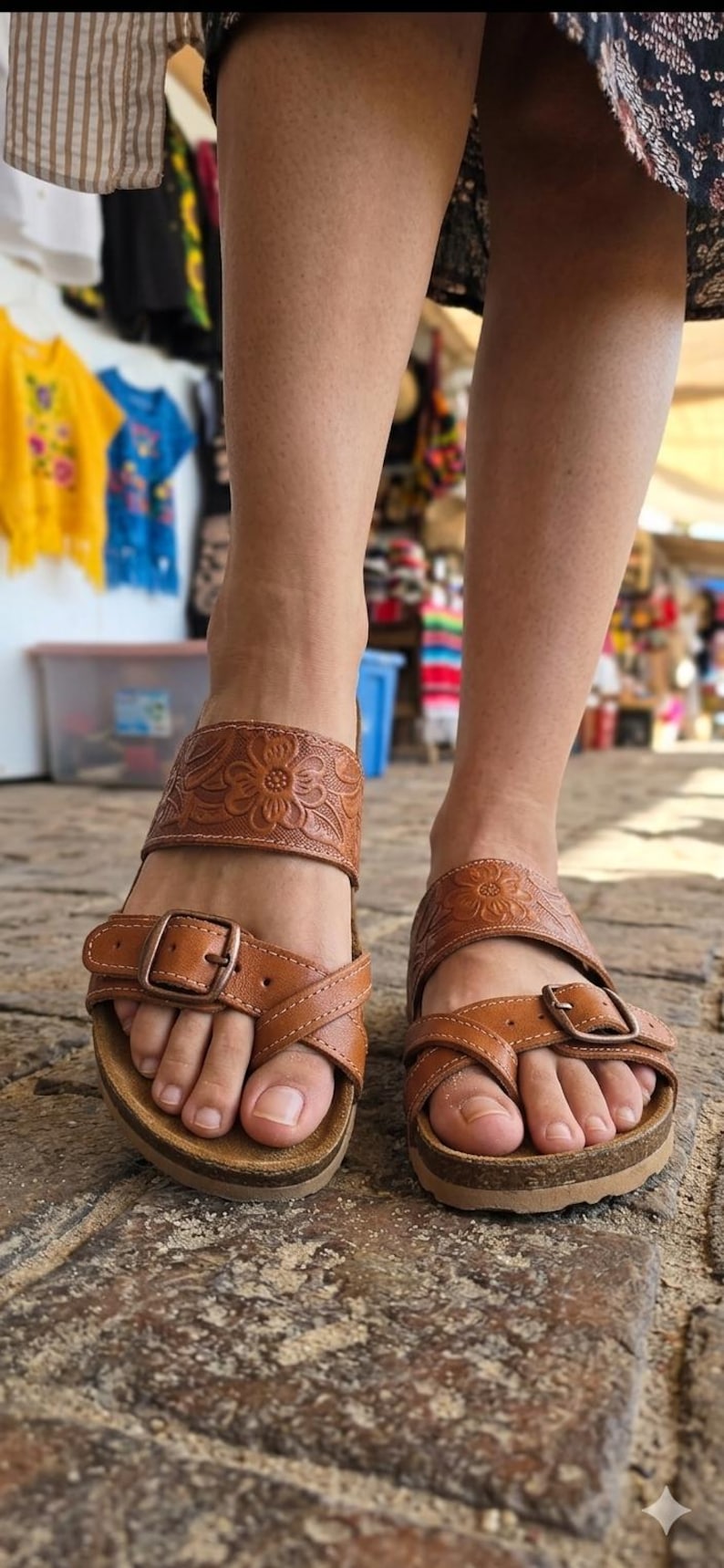 Pu&ograve; includere: Sandali in pelle marrone con dettagli floreali in rilievo sui cinturini. I sandali hanno un plantare in sughero e fibbie regolabili. L'immagine mostra i sandali ai piedi di una persona, con uno sfondo sfocato.