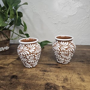 May include: Two small, brown ceramic vases with white floral patterns. The vases have a white rim and are displayed on a wooden surface. The image also includes a green plant and the text "Mexicanita Handmade".