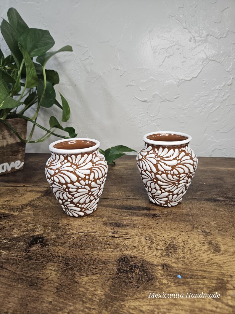 May include: Two small, brown ceramic vases with white floral patterns. The vases have a white rim and are displayed on a wooden surface. The image also includes a green plant and the text "Mexicanita Handmade".