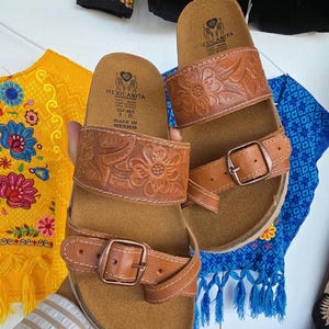 Pu&ograve; includere: Sandali in pelle marrone con cinturini in rilievo floreale e chiusure con fibbia. I sandali hanno un plantare in sughero e sono etichettati "MEXICANITA HANDMADE". Taglia 8.25.