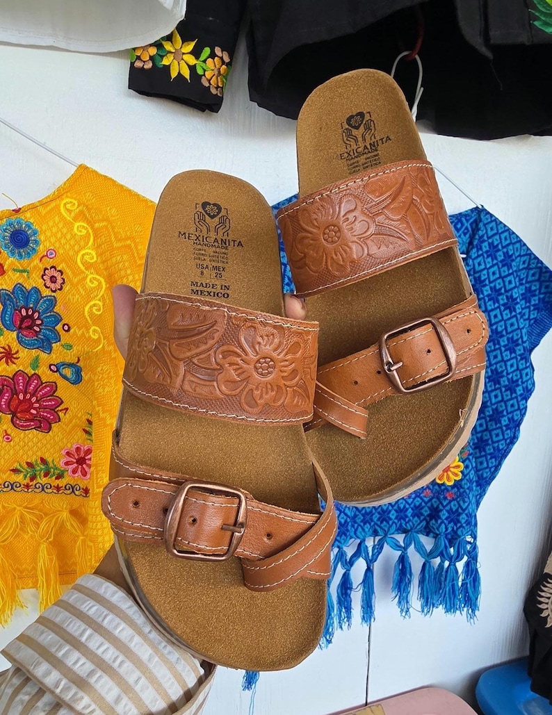 Pu&ograve; includere: Sandali in pelle marrone con cinturini in rilievo floreale e chiusure con fibbia. I sandali hanno un plantare in sughero e sono etichettati "MEXICANITA HANDMADE". Taglia 8.25.