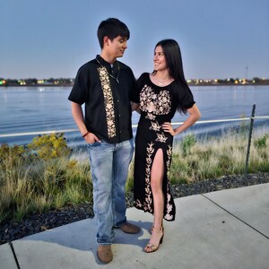 May include: A couple standing on a sidewalk by a body of water. The woman is wearing a black dress with embroidery and the man is wearing a black shirt with embroidery. They are both looking at each other.