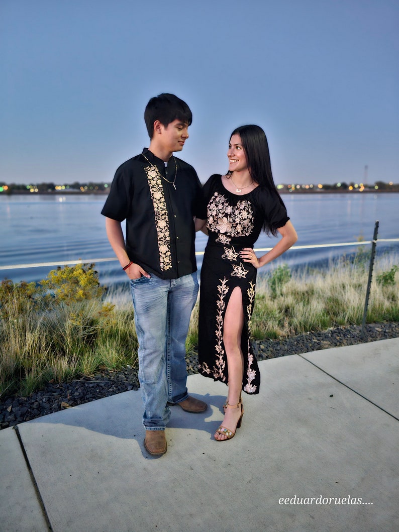 May include: A couple standing on a sidewalk by a body of water. The woman is wearing a black dress with embroidery and the man is wearing a black shirt with embroidery. They are both looking at each other.