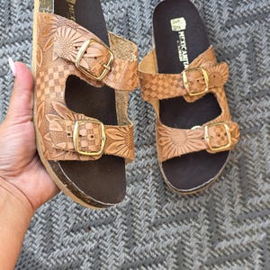Op de afbeelding: Een paar lichtbruine leren sandalen met twee verstelbare bandjes met goudkleurige gespen. Het leer is voorzien van een reli&euml;f met een zonnebloem- en dambordpatroon. Het voetbed is donkerbruin en de merknaam MEXICANITA is in het wit gedrukt.