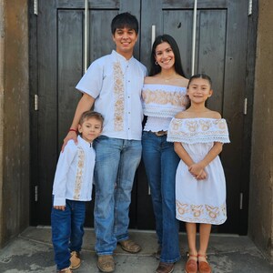 Puede incluir: Una familia de cuatro personas vestidas con camisas y vestidos blancos bordados. El padre lleva pantalones vaqueros azules y zapatos marrones. La madre lleva pantalones vaqueros azules y sandalias marrones. Los dos ni&ntilde;os llevan vestidos blancos y sandalias marrones. La familia est&aacute; de pie frente a una puerta de madera.