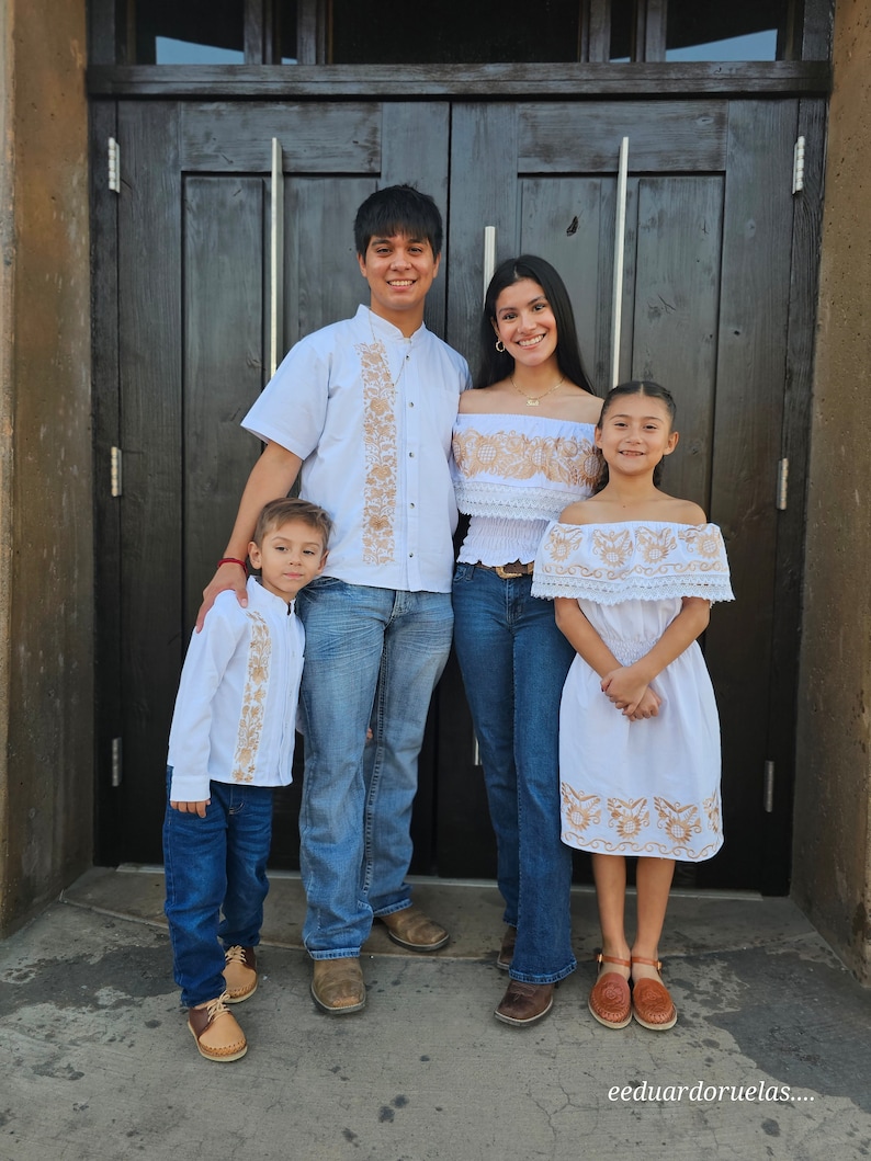 Puede incluir: Una familia de cuatro personas vestidas con camisas y vestidos blancos bordados. El padre lleva pantalones vaqueros azules y zapatos marrones. La madre lleva pantalones vaqueros azules y sandalias marrones. Los dos ni&ntilde;os llevan vestidos blancos y sandalias marrones. La familia est&aacute; de pie frente a una puerta de madera.