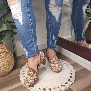 May include: Brown leather shoes with white floral embroidery. The shoes have a block heel and a cutout design. The person is wearing blue denim jeans with distressed details. The shoes are on a white, round rug with tassels. The text "Mexicanita Handmade" is visible.