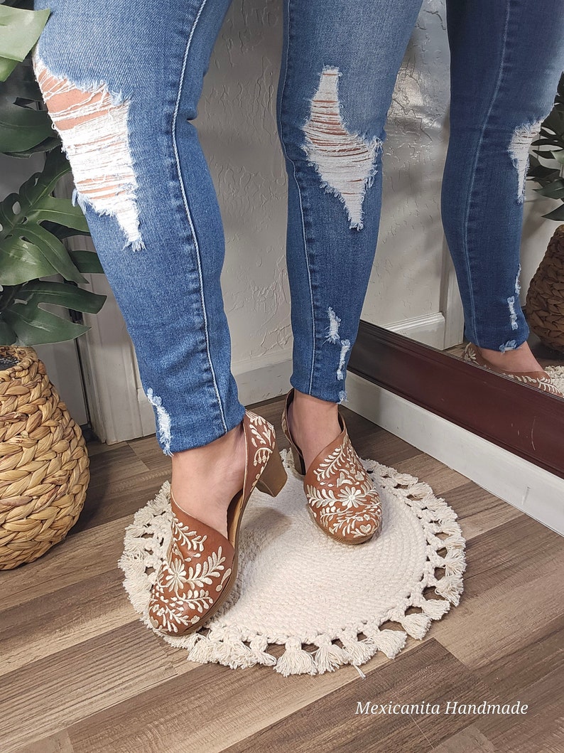 May include: Brown leather shoes with white floral embroidery. The shoes have a block heel and a cutout design. The person is wearing blue denim jeans with distressed details. The shoes are on a white, round rug with tassels. The text "Mexicanita Handmade" is visible.