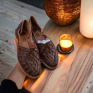 Puede incluir: Un par de sandalias de cuero marr&oacute;n con correas tejidas. Las sandalias est&aacute;n sobre una superficie de madera con una vela y un cuenco decorativo en el fondo.