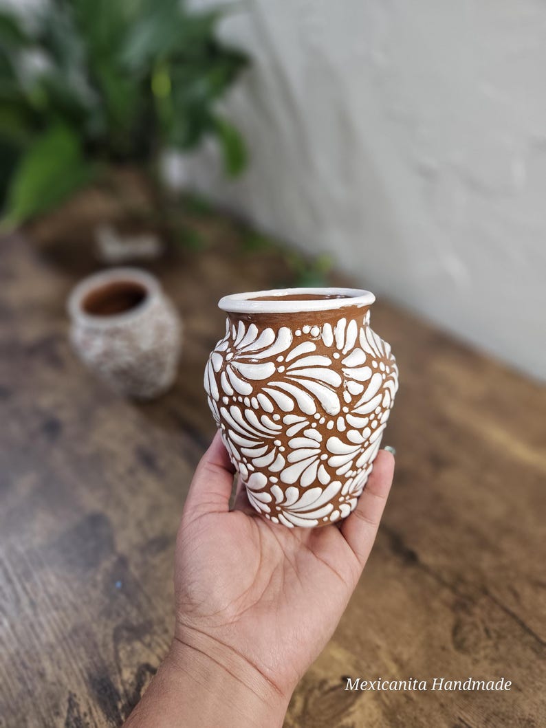 May include: A small, brown ceramic vase with a white floral pattern. The vase is held in a hand and has a rounded shape with a wide opening. The text "Mexicanita Handmade" is visible.