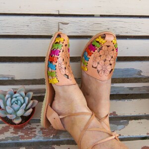 Puede incluir: Un par de sandalias de cuero marr&oacute;n con bordados florales de colores. Las sandalias tienen correas de tobillo y se usan en los pies de una persona.