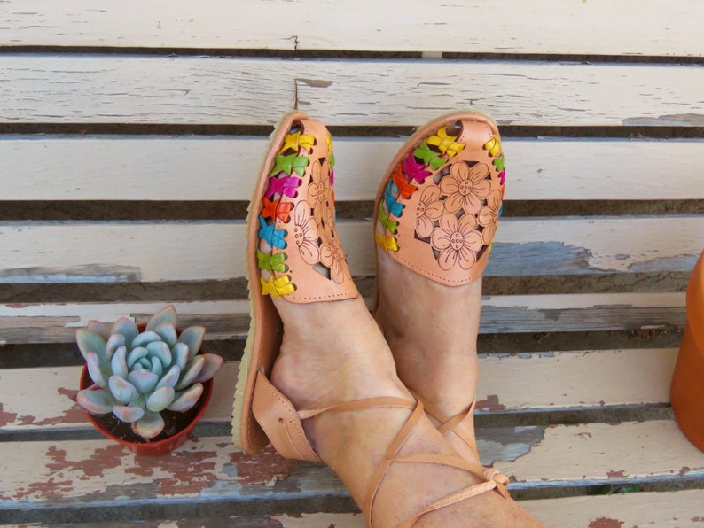 Puede incluir: Un par de sandalias de cuero marr&oacute;n con bordados florales de colores. Las sandalias tienen correas de tobillo y se usan en los pies de una persona.