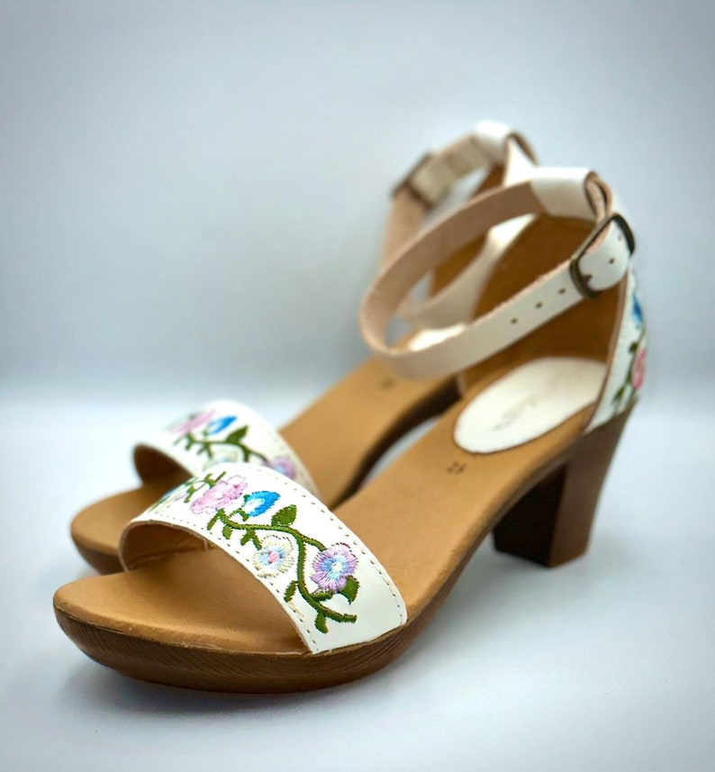 Puede incluir: Sandalias de cuero blanco con un dise&ntilde;o de bordado floral. Las sandalias tienen un tac&oacute;n de madera marr&oacute;n y una correa de tobillo con cierre de hebilla.
