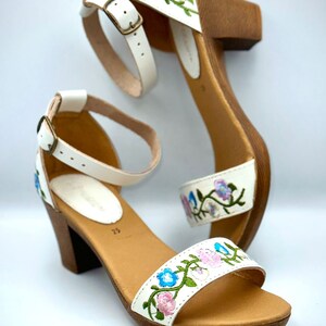 Puede incluir: Sandalias blancas de cuero con tac&oacute;n y detalles florales bordados en rosa, azul y verde. Las sandalias tienen una plataforma de madera y una correa de tobillo con cierre de hebilla.