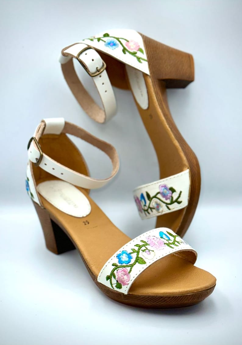 Puede incluir: Sandalias blancas de cuero con tac&oacute;n y detalles florales bordados en rosa, azul y verde. Las sandalias tienen una plataforma de madera y una correa de tobillo con cierre de hebilla.