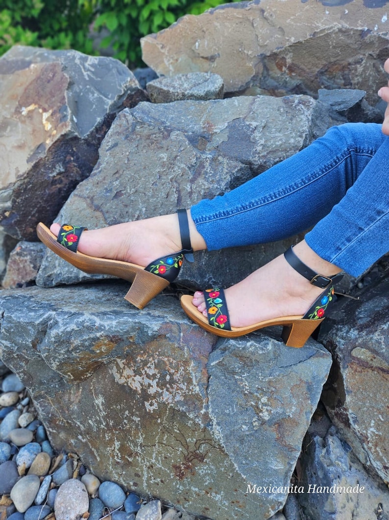 May include: Black leather sandals with a wooden heel and colorful floral embroidery. The sandals are worn by a person sitting on rocks.
