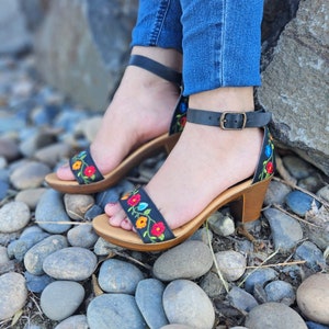 May include: Black leather sandals with a floral embroidery design. The sandals have a block heel and an ankle strap with a buckle closure.