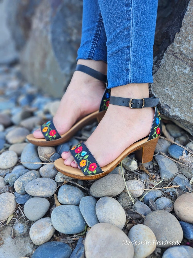 May include: Black leather sandals with a floral embroidery design. The sandals have a block heel and an ankle strap with a buckle closure.
