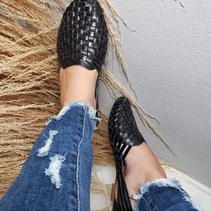 May include: A pair of black woven leather sandals with a cut-out design. The sandals are worn by a person wearing blue jeans.