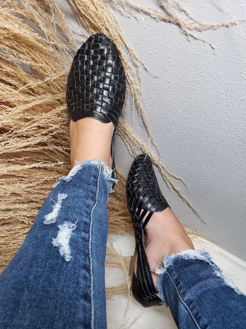 May include: A pair of black woven leather sandals with a cut-out design. The sandals are worn by a person wearing blue jeans.