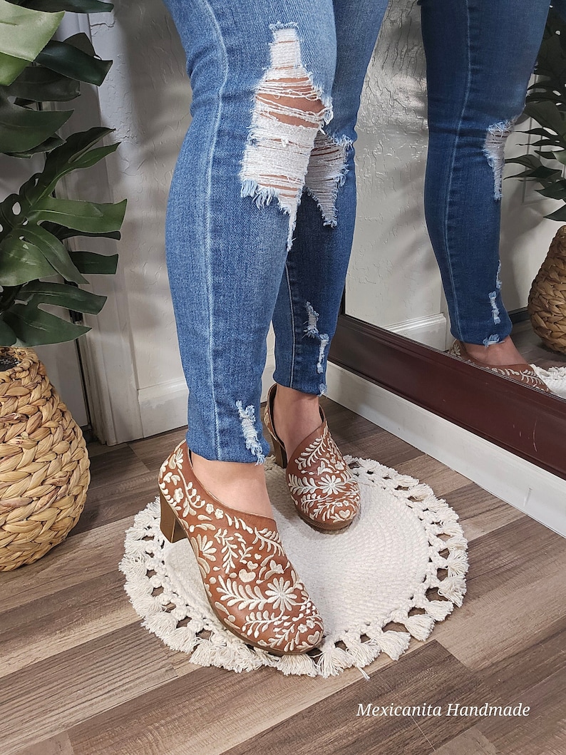 May include: Brown leather shoes with white floral embroidery. The shoes have a block heel and a low-cut design. The shoes are worn with blue jeans. The text "Mexicanita Handmade" is visible in the image.