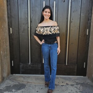 May include: A woman wearing a black off-the-shoulder top with embroidered floral details in a beige colour. She is wearing blue jeans and brown boots. The top has a fitted bodice and a flowy, slightly ruffled hem. The woman is standing in front of a wooden door.