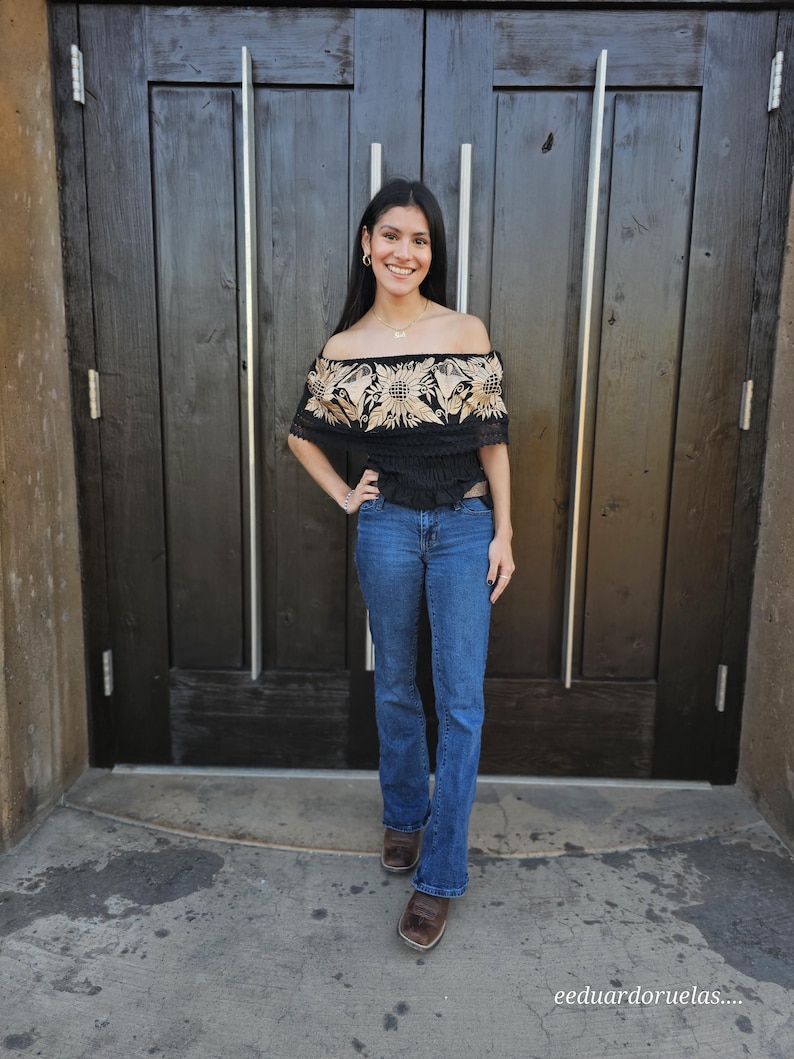 May include: A woman wearing a black off-the-shoulder top with embroidered floral details in a beige colour. She is wearing blue jeans and brown boots. The top has a fitted bodice and a flowy, slightly ruffled hem. The woman is standing in front of a wooden door.