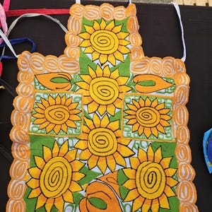 Puede incluir: Un delantal de cocina blanco con un dise&ntilde;o de girasoles verdes y amarillos. El delantal tiene un borde festoneado con un ribete amarillo. El texto "Mexicanita Handmade" es visible en la esquina inferior derecha.