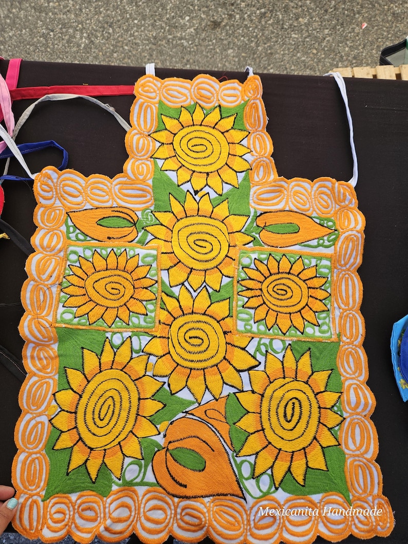 Puede incluir: Un delantal de cocina blanco con un dise&ntilde;o de girasoles verdes y amarillos. El delantal tiene un borde festoneado con un ribete amarillo. El texto "Mexicanita Handmade" es visible en la esquina inferior derecha.