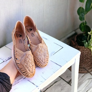 Puede incluir: Un par de sandalias de cuero marr&oacute;n con correas tejidas. Las sandalias est&aacute;n sobre una superficie de madera blanca.