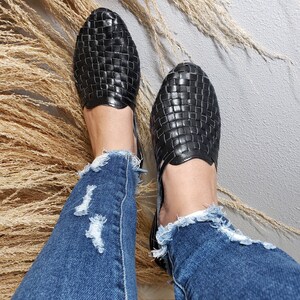 May include: A pair of black woven leather sandals with a criss-cross pattern. The sandals are worn by a person wearing blue jeans.