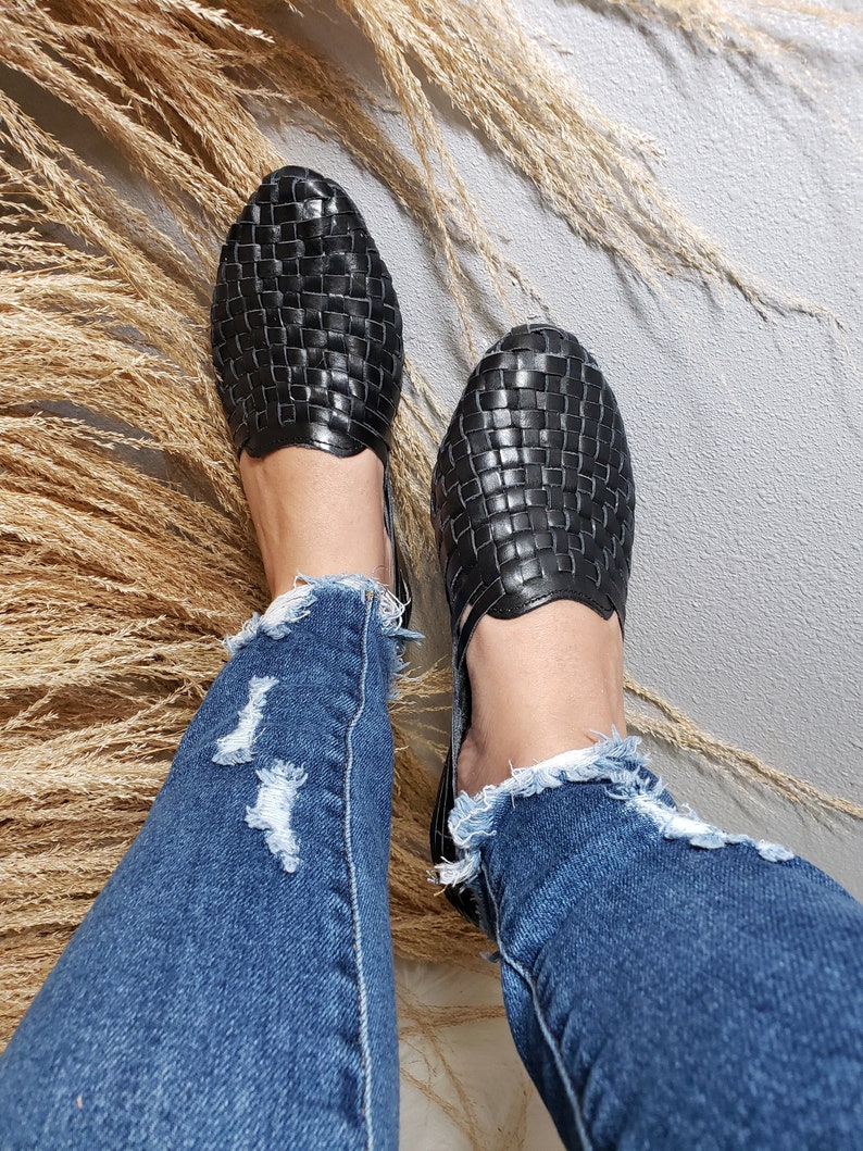 May include: A pair of black woven leather sandals with a criss-cross pattern. The sandals are worn by a person wearing blue jeans.